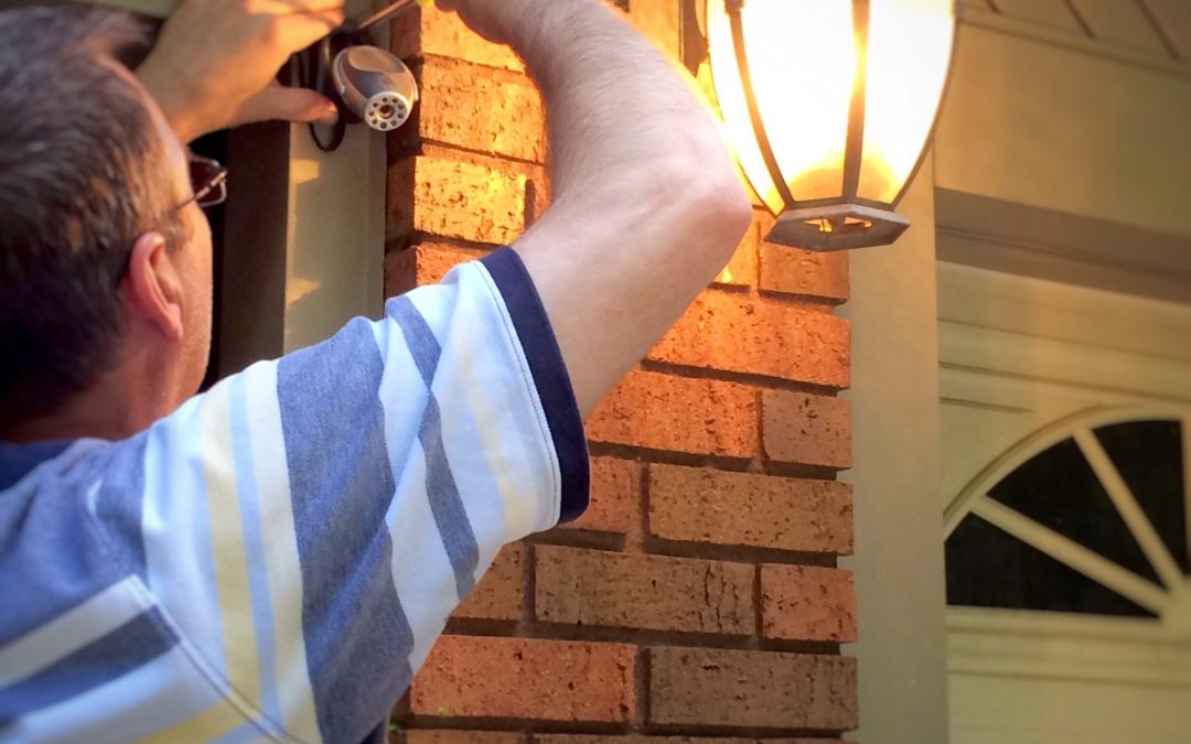 An older man installs a camera outside of his brick home. A porch light is lit casting a warm glow.